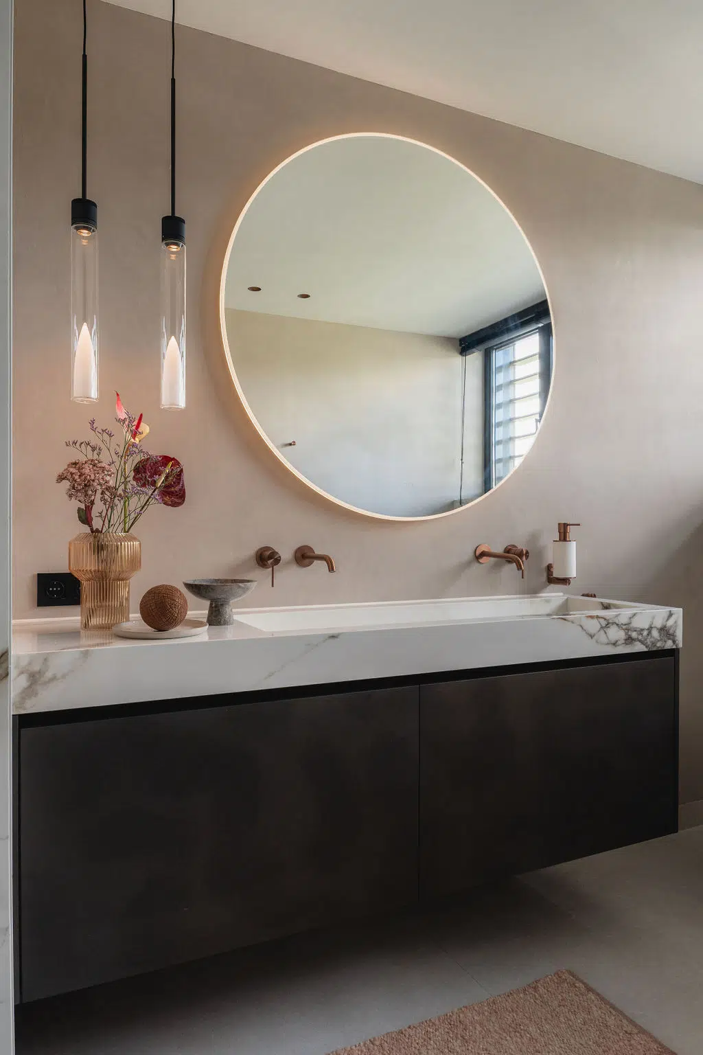 Impressie van een design badkamer met een lichte uitstraling. De badkamer heeft een luxe bad met vrijstaande kraan in koperen kleur, geplaatst onder het raam en tegen een marmerlook tegel. Naast het bad zien we een royaal meubel met grote wastafel, een grote ronde spiegel en wandkranen in de kleur koper.