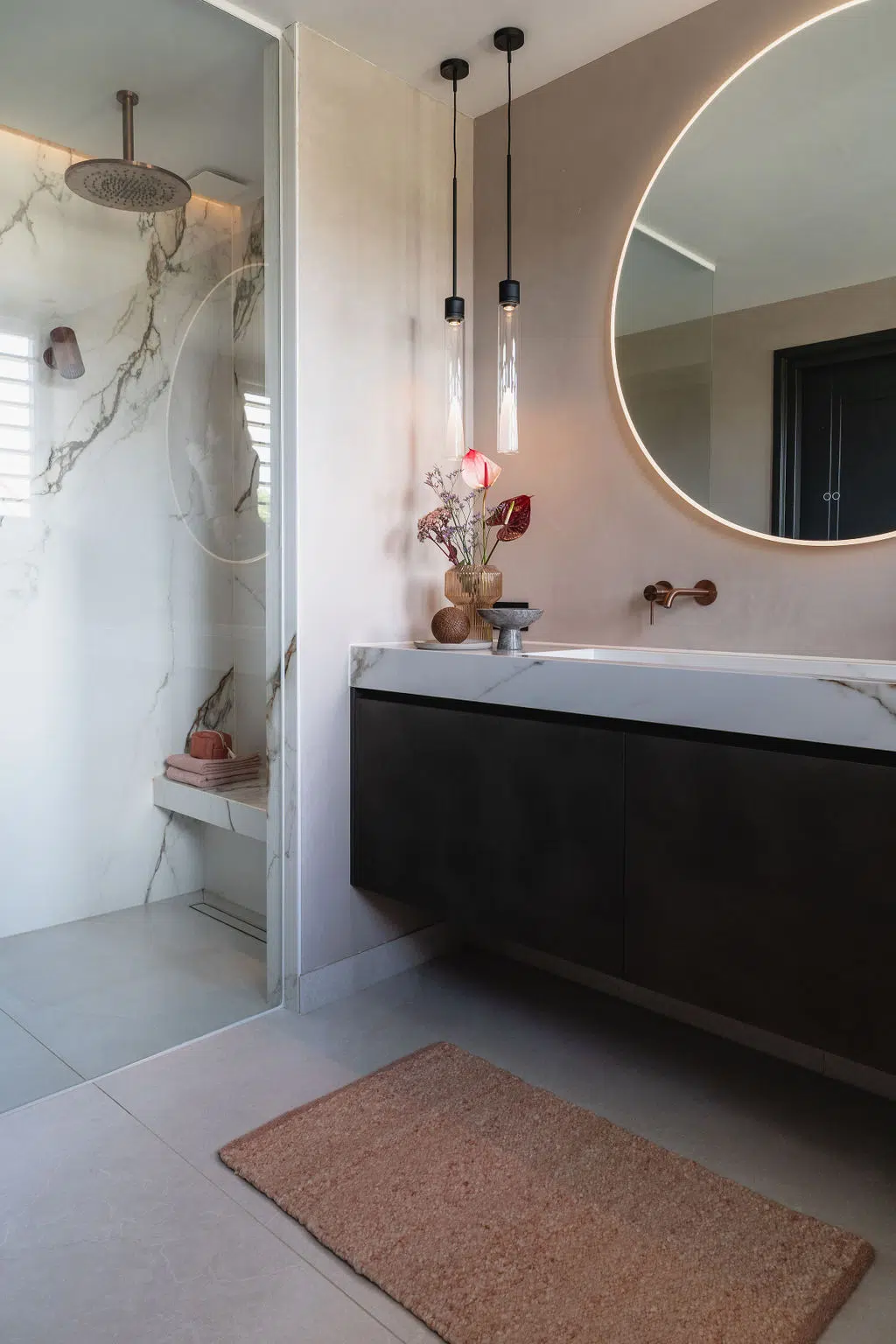 Impressie van een design badkamer met een lichte uitstraling. De badkamer heeft een royaal meubel met grote wastafel, een grote ronde spiegel en wandkranen in de kleur koper. Naast het badkamermeubel zien we een walk-in-douche met regendouche in dezelfde kleur koper.