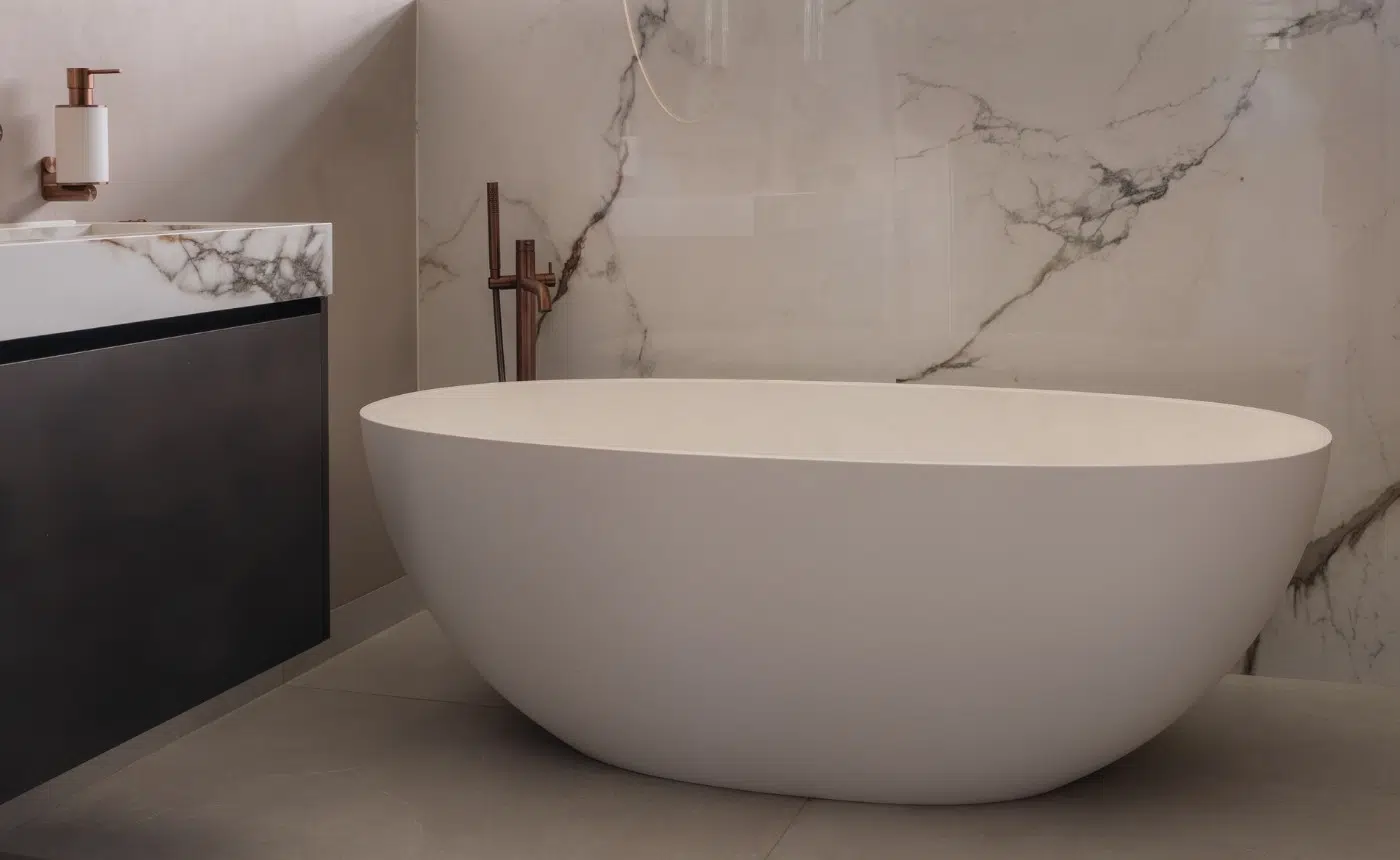 Impressie van een design badkamer met een luxe bad met vrijstaande kraan in koperen kleur, geplaatst onder het raam en tegen een marmerlook tegel.