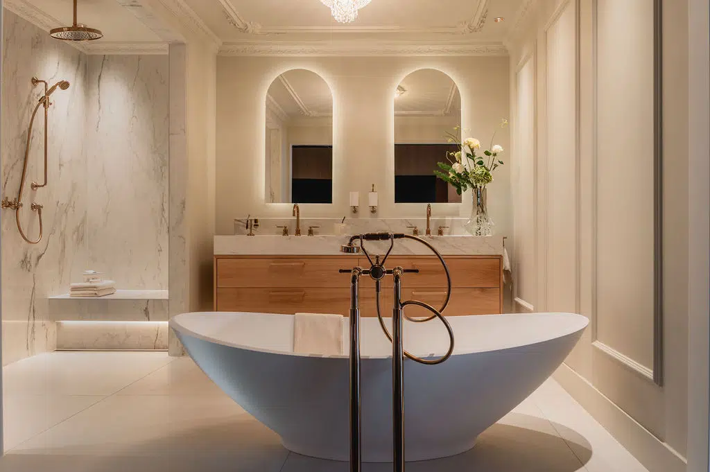 Impressie van een klassieke badkamer, voorzien van sierlijsten, een luxe ligbad, warme accenten, een maatwerk badkamermeubel en marmeren slabs.