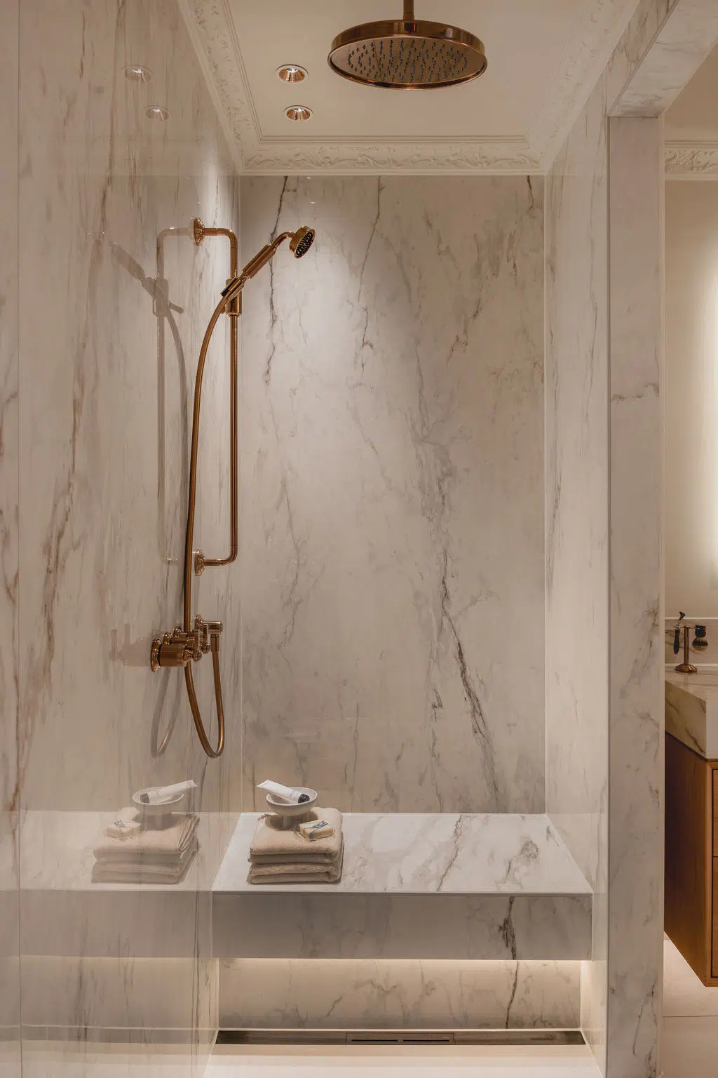 Douche met glazen deur, voorzien van een Gessi doucheset en scharnieren in de kleur messing. De douche heeft marmeren slabs en een bankje.