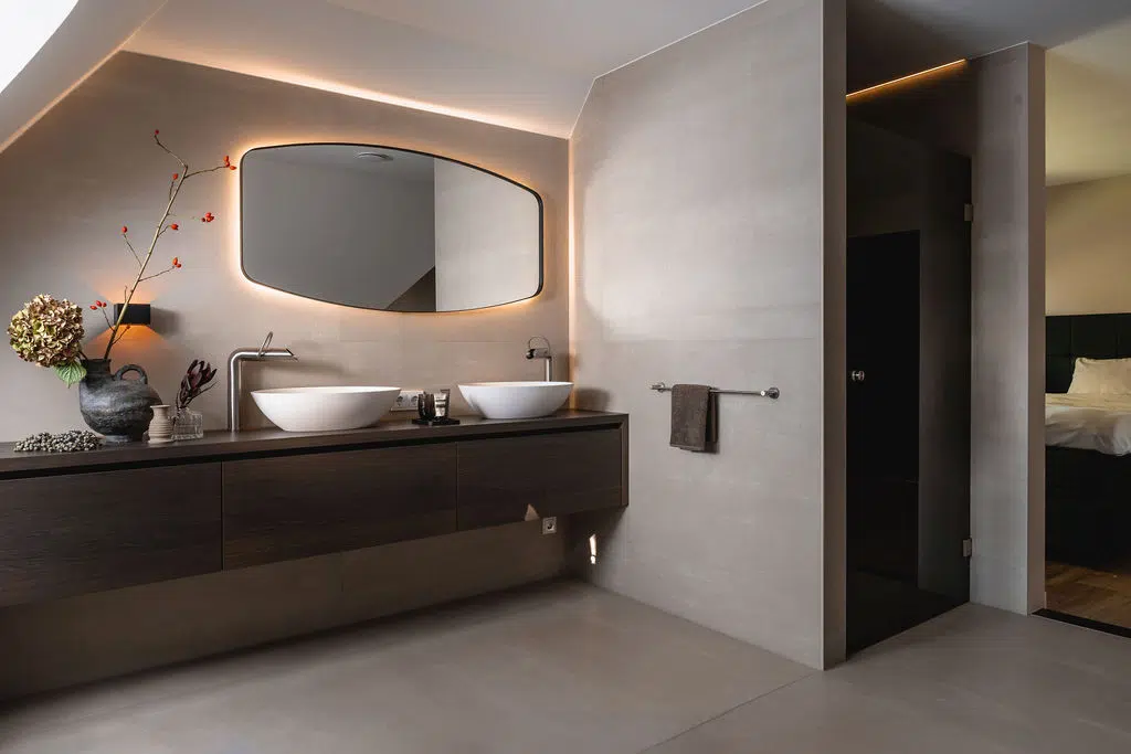 Impressie van een landelijke badkamer met grote wand- en vloertegels, een maatwerk badkamermeubel van hout, twee luxe waskommen en een grote spiegel met verlichting.