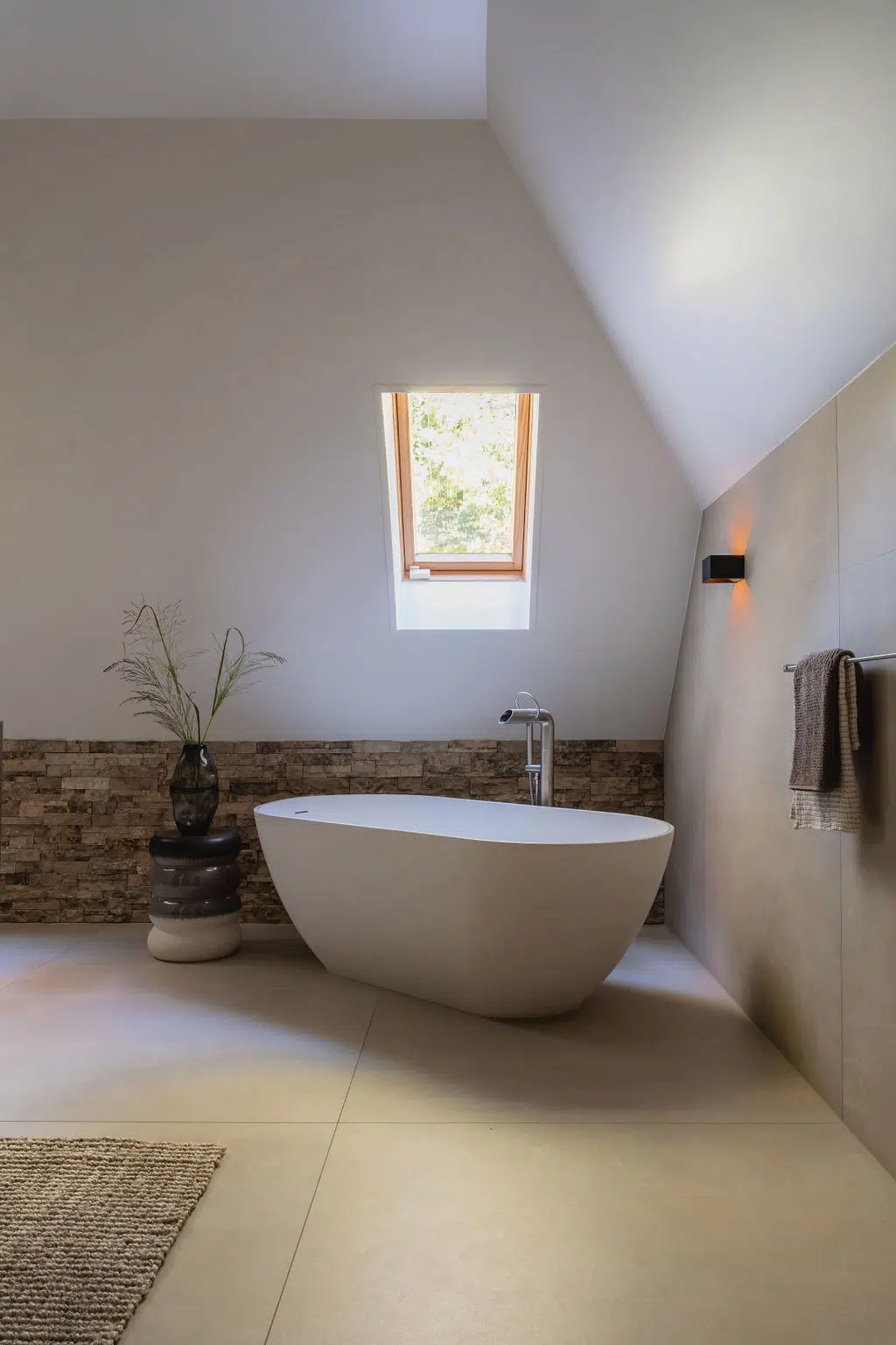 Impressie van een landelijke badkamer met grote vloertegels en wandtegels. We zien een vrijstaand bad onder een dakraam, met daarachter een wandje met natuursteen strips.