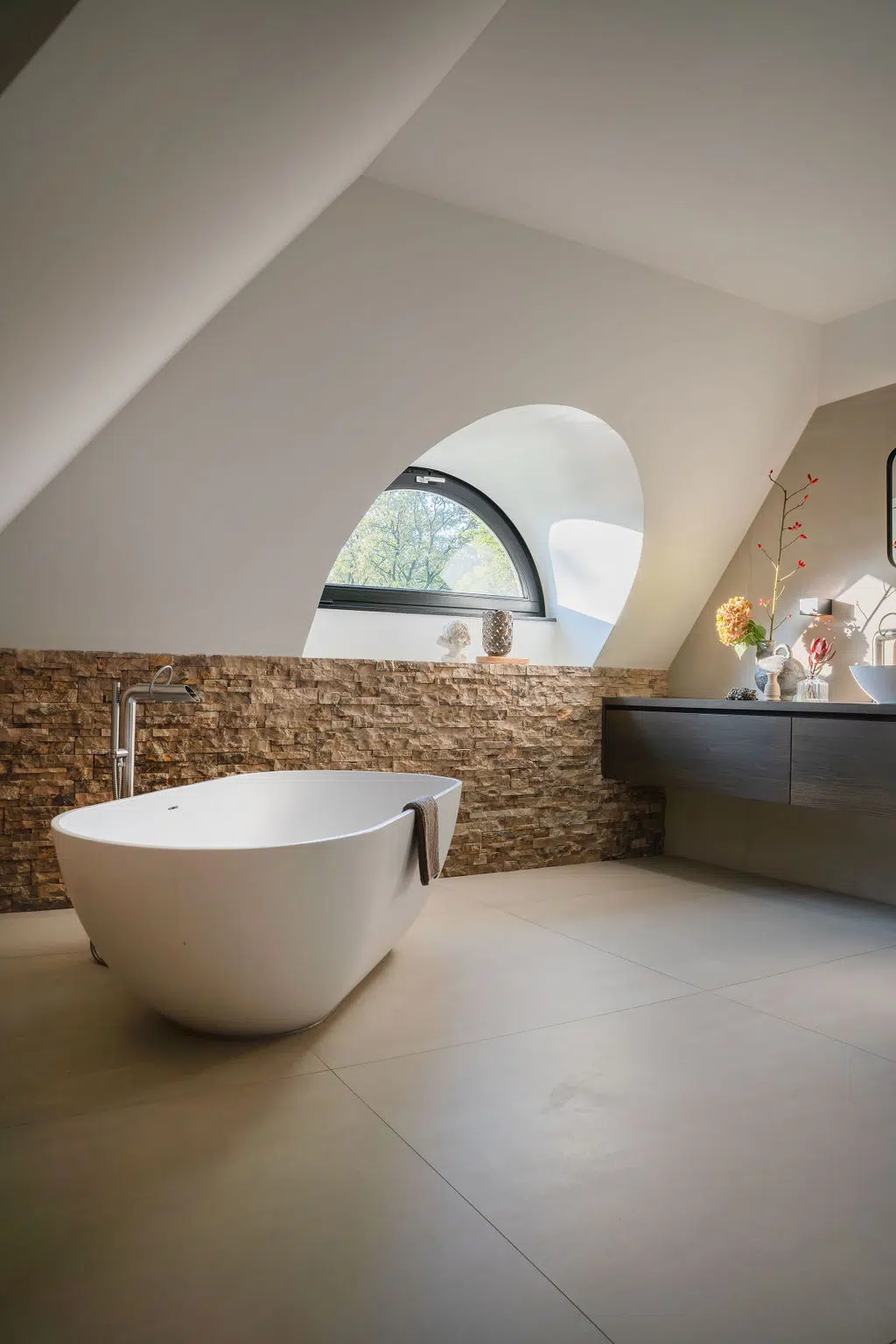 Impressie van een landelijke badkamer met riante lichtinval vanuit een stijlvol dakkapel. De badkamer heeft grote wand- en vloertegels, een wand met natuursteen strips, een maatwerk badkamermeubel van hout, een vrijstaand bad en een grote spiegel met verlichting.