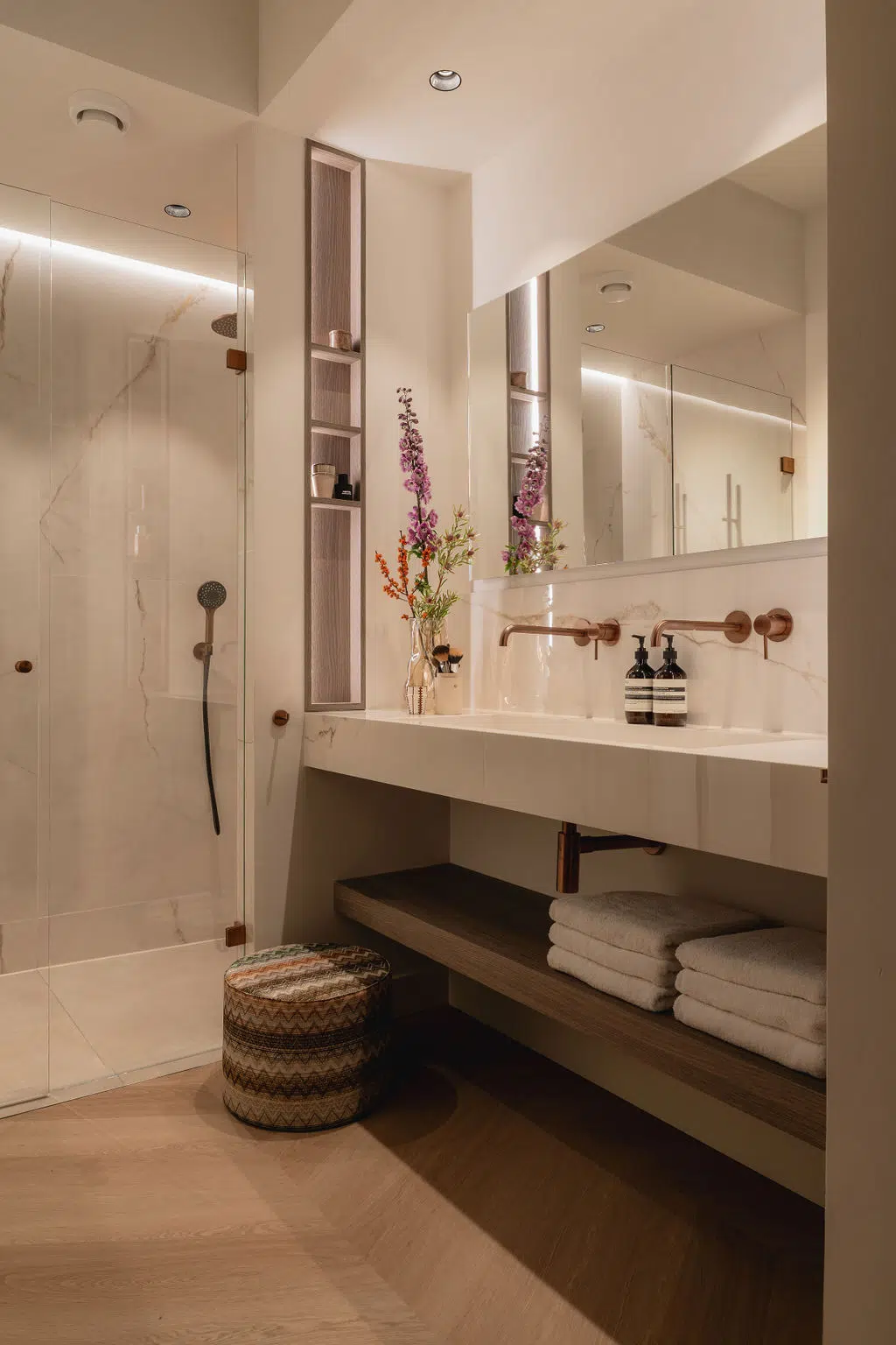 Impressie van een kleine badkamer met een donker hout badkamermeubel, zwevende badkamerkast, koper kranen, een grote spiegel en extra sfeer verlichting bij de douche.