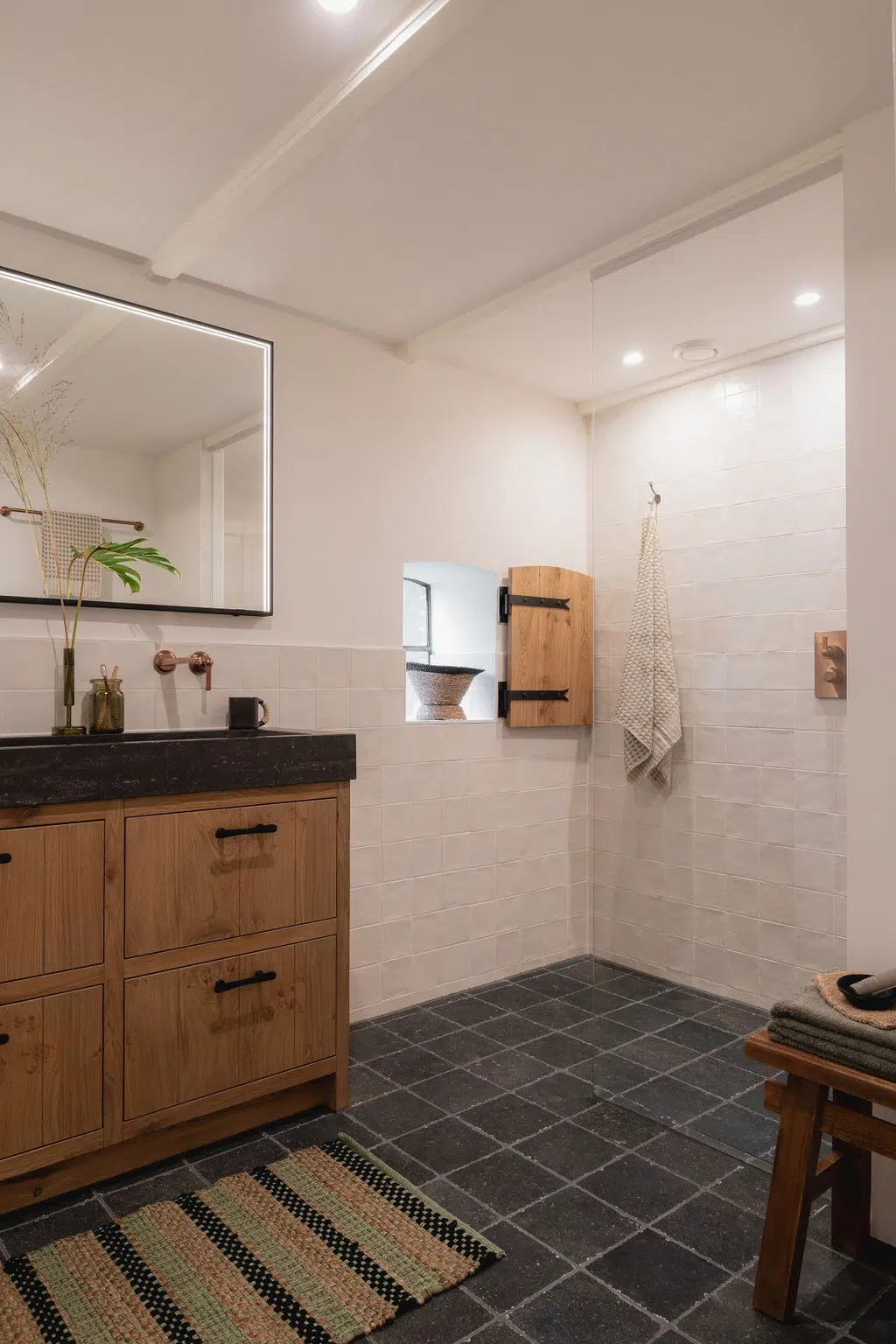 Impressie van een landelijke badkamer met vierkante tegels: witte wandtegels en grijze vloertegels. Ook zien we Gessi kranen in geborsteld koper en een walk-in douche.