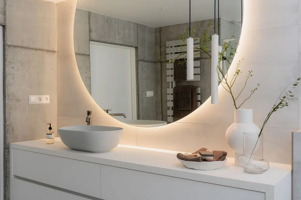 Impressie van een moderne badkamer met grijze tegels, een grote wastafel met waskom en kraan in zwart chroom, en een halfronde spiegel met verlichting.