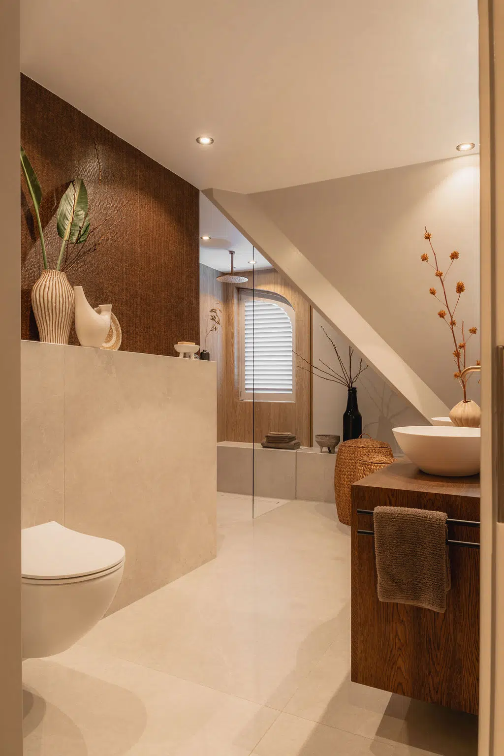Impressie van een Japandi badkamer met schuine wand. We zien een basis van warme badkamer kleuren, een lambrisering met hangend toilet, een nis met unieke ronding erin en behang.