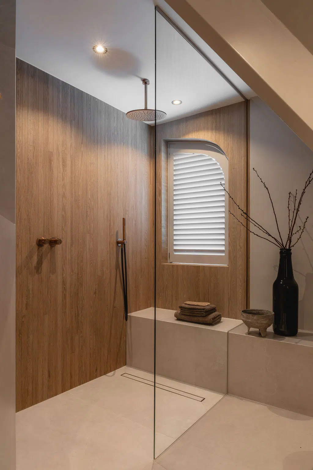 Impressie van een doucheruimte met maatwerk glazen douchewand, houtlook wandtegels, bankje en Gessi doucheset met handdouche en stortdouche. De douche heeft een nis met een unieke ronding aan de bovenkant.