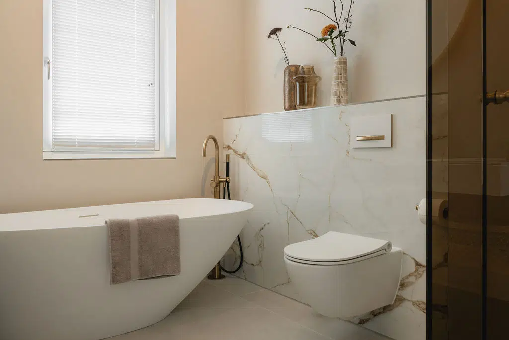 Impressie van een luxe badkamer met vrijstaand bad en een lambrisering met hangend toilet. De wand is voorzien van een marmeren slab met een gouden ader; de kleur die we ook terugzien bij de vrijstaande badkraan.