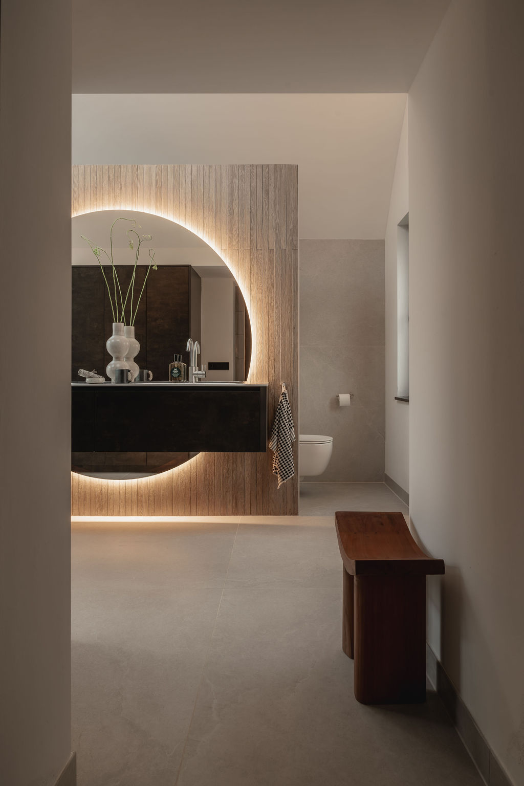 Binnenkomst bij een moderne badkamer met donker badkamermeubel, bevestigd aan een wand met houtlook tegels. Achter het meubel loopt een royale ronde spiegel met verlichting. De spiegel steekt zowel aan de onderkant als bovenkant van het meubel uit.