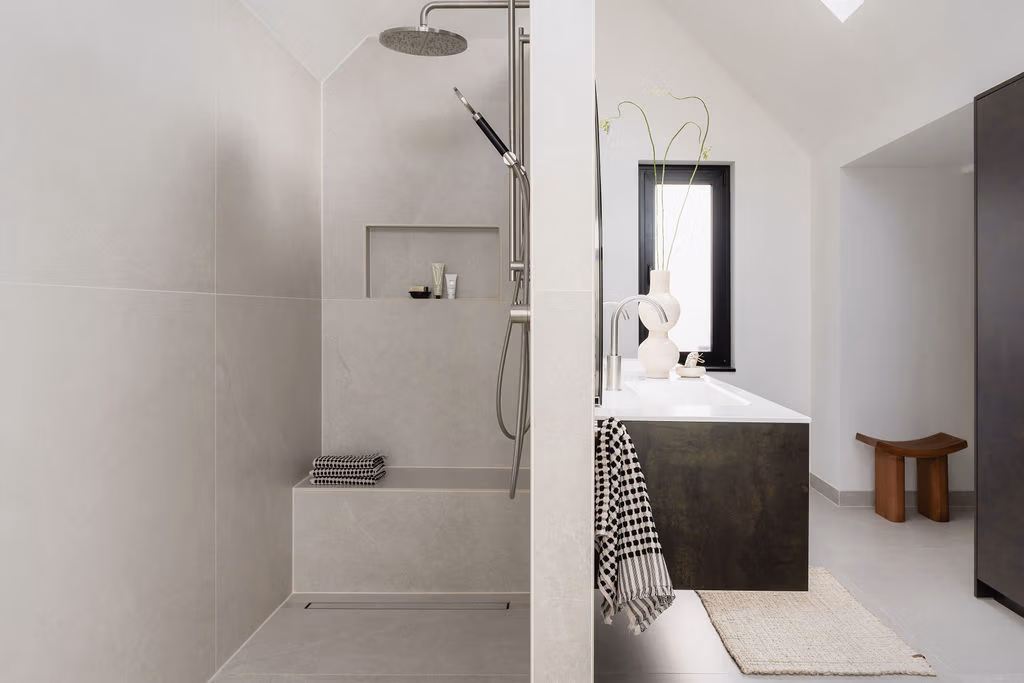 Impressie van een moderne badkamer met tussenwand. Aan de ene zijde zien we een luxe doucheruimte met twee regendouches. Aan de andere zijde zien we een houten badkamermeubel met RVS kranen.
