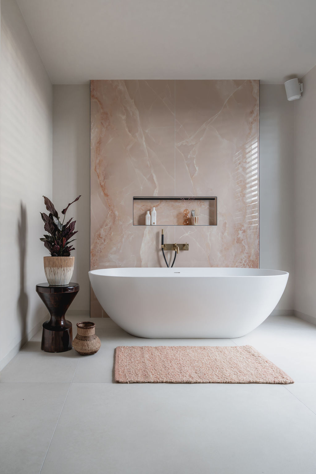 Impressie van een luxe wellness badkamer, voorzien van vrijstaand bad en een kraan in geborsteld messing. Het bad staat voor een wand met zachtroze marmerlook slabs.