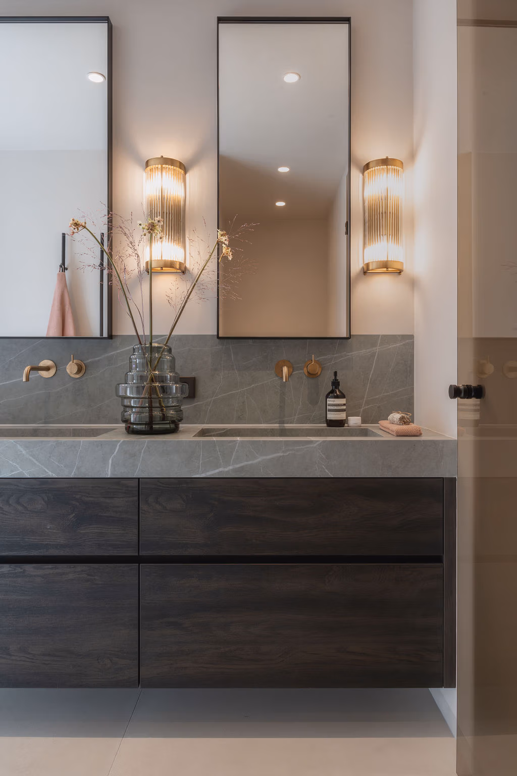 Impressie van een luxe badkamermeubel met donker houten onderkast, een groengrijze marmerlook wastafel, kranen in geborsteld messing en luxe spiegels.